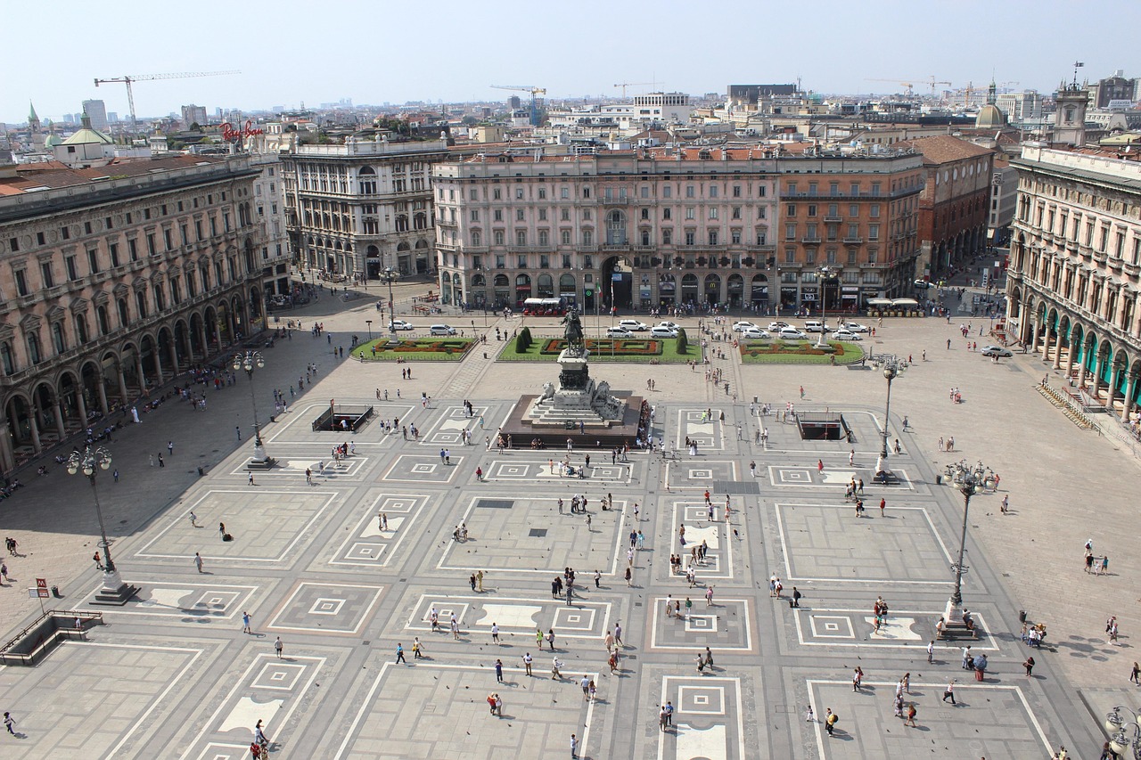 Piazza affollata con persone che si fermano, monumenti storici sullo sfondo.
